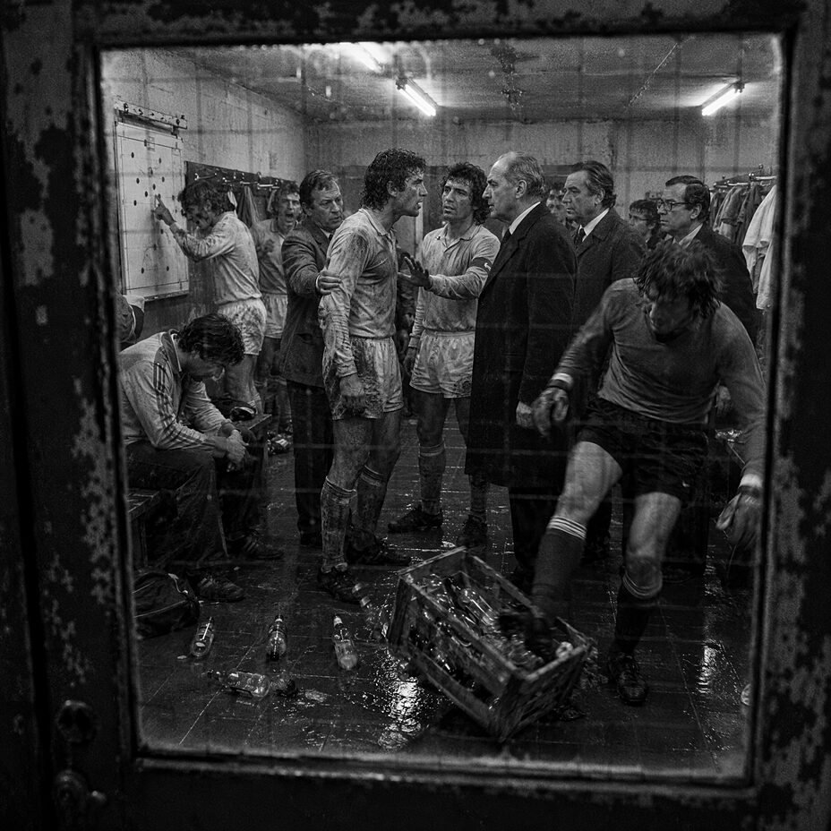 A national team dressing room at half-time in 1978, players in muddy kit confronting officials in overcoats, an overturned water crate on the floor